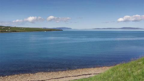 A large expanse of water. Some small hills in the background and a small pebble beach in the foreground. A blue sky with small, light clouds