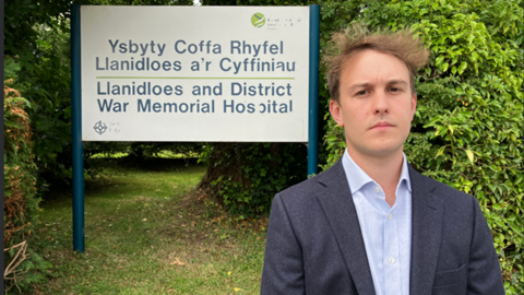 County Councillor Glyn Preston looking into the camera stood in front of Llanidloes Hospital sign. 