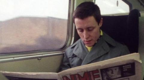 A man reads an NME newspaper on a train.