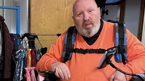 Mick Lloyd looking at the camera and wearing an orange jumper with his elbows leaning on his standing wheelchair. 