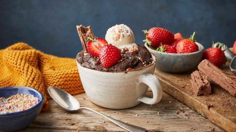 A chocolate mug cake with a scoop of icing, sprinkles, strawberries and a flaky chocolate bar on top