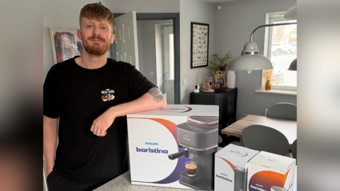 A man standing in a kitchen beside a boxed Philips “Baristina” coffee machine, with a dining table, chairs, hanging lamp and houseplants visible in the background.