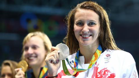 A woman with damp brown hair smiling as she holds up a silver Olympic medal. She wears a white Team GB jersey bearing the Olympic rings logo.