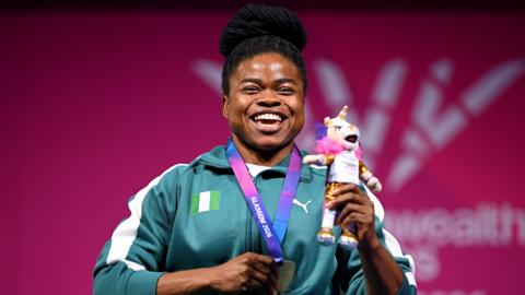 Esther Nworgu smiles broadly during the medal ceremony for the lightweight para-powerlifting at the 2026 Commonwealth Games. Visible from the waist up, she wears a green and white Nigerian tracksuit and uses her right hand to clutch her gold medal, which is on a purple and pink ribbon around her neck, and her left to hold up a cuddly toy Games mascot. Blurred in the background is a red board with a white Glasgow 2026 logo on it