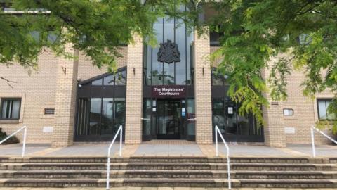 Great Yarmouth Magistrates' Court house, a yellow brick building with a colonnaded entrance and brown-framed windows. There are steps and hand rails approaching the entrance, and green trees overhanging in the foreground of the image.