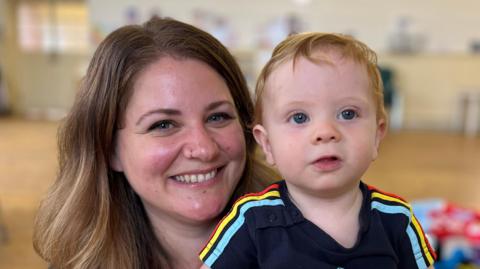 A woman with long brown hair grins while holding her baby. He has short ginger hair and wears a navy top with red, yellow and blue stripes down the sleeves.
