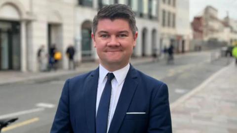 A picture of Sam smiling at the camera. He has black hair and is wearing a white suit shirt, navy tie and a navy blazer. 