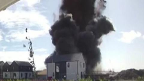 A plume of smoke rises above a house after a controlled explosio