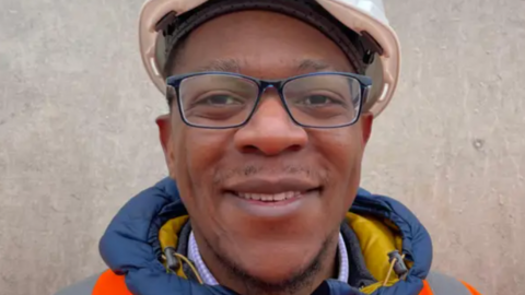 A head shot of a Severn Trent worker in a white hard hat and an orange hi-vis jacket. The man is smiling and standing in front of a wall.