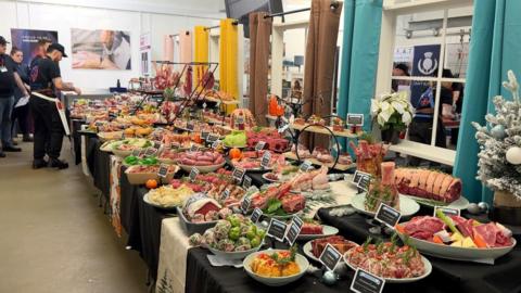 Ornately displayed meat dishes including roasting cuts tied with string, stir fry and curry dishes and sausages one a 15-foot table. There are a group of people in black t-shirts to the left. 