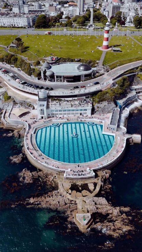 A drone shot of a large seaside pool. There is a lighthouse in the background.