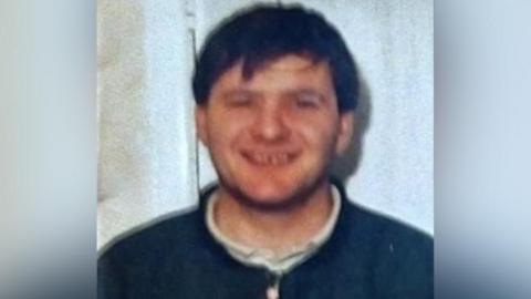 An old photo of Paul Thompson. It is slightly out of focus. He is smiling at the camera. He has dark hair which falls over his forehead.  He is wearing a dark blue top with a white collared shirt underneath, 