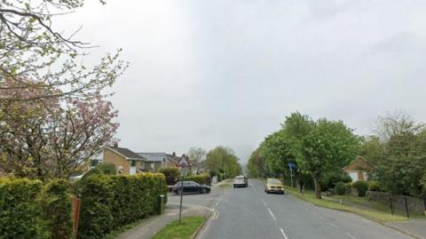 A  road lined with trees and houses. There are two cars on the main road with another car pulling out at the junction with the road. 