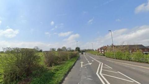 A road with a field to the left hand side and housing to the right hand side. There is a traffic filter lane in the centre of the road, and arrows directing traffic.