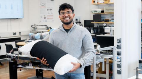 PhD researcher Malindu Ehelagasthenna wearing a grey jumper and glasses, holding a pillow and smiling stood in a lab at Nottingham Trent University.