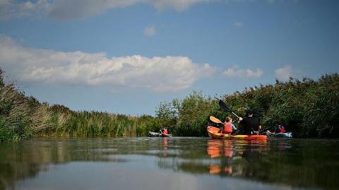 Three kayaks on a still river with high grass on its banks. The nearest kayak contains one adult in a hat and a child wearing a lifejacket. The sky is blue with white, fluffy clouds.