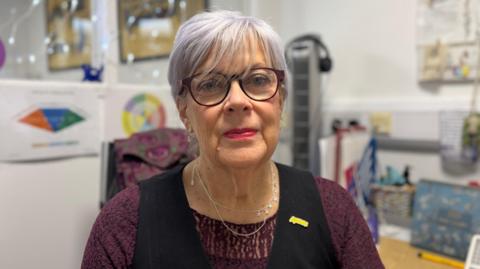 A close-up image of Cherry Cantrell sitting at the desk in her office. She is an older woman, with a purple hue in her short hair. She is wearing  apair of circular glasses, a silver necklace, as well as a purple lace top and black waistcoat. She has a yellow 'Aware NI' pin on her lapel. She is looking at the camera and smiling. Behind her are posters and lights on the wall of her office, but they are blurred.
