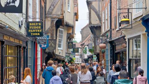 View along The Shambles in York, a narrow medieval street with timber-framed buildings housing shops and cafes. The scene shows the busy pedestrian area that retains its characteristic overhanging upper stories and historic layout.