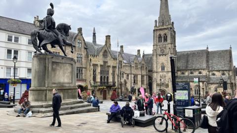 A town square on a grey day with lots of people milling about. There is a large statue of a knight on horseback in the foreground, while an old sandy coloured church with large steeple is at the rear of the square.