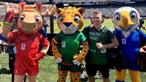 A man in a green photographers bib, with a camera over his shoulder, is standing between three football mascots, alongside the pitch in a stadium