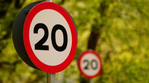 A 20mph sign with another behind it. It's a circular sign with red borders and black text reading '20'.
