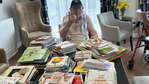 Stan Woolley, 100, wearing a black and green baseball cap with a military logo on the front and a beige polo neck, sits in front of a dining table laden with hundreds of birthday cards of all different colours. 