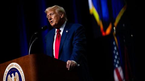 Donald Trump speaks into a microphone while standing next to a lectern. He is wearing a suit and red tie.