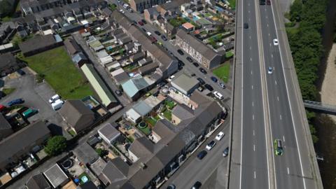 A motorway with cars and vans on it running over the top of a row of terraced houses, seen from overhead