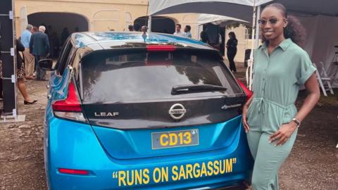 Biologist Shanika Spencer poses next to a blue Nissan Leaf car on which the sentence "Runs on Sargassum" is printed. 