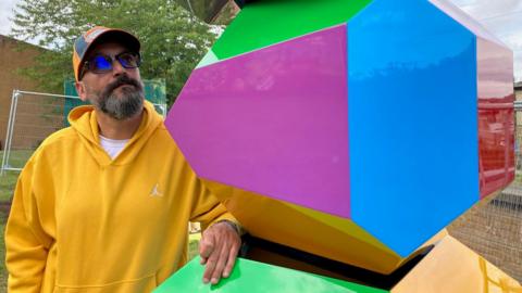 A man in a yellow hooded top and baseball cap looking at a multi-coloured block.
