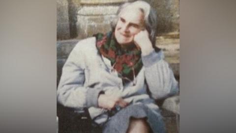 A grainy scan of a portrait photo of Mary Body sitting on a bench. She has grey shoulder-length hair and is wearing a light grey jacket and medium grey skirt along with a black, red and green floral scarf. She is also wearing glasses on a chain around her neck. She is looking off to the right of the camera, is smiling and has her left cheek propped up on her left knuckles. It can just about be made out that the wooden bench in directly in front of a stone building.