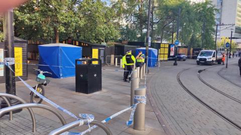 Image shows Piccadilly Gardens in Manchester, where police tape has been wrapped around several bollards and lampposts. Three police officers stand in the background. 