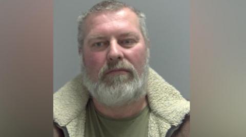 Trevor New-Watts is looking at the camera in a police custody shot. He has short greying hair and facial hair. He is wearing a green top and a brown jacket with a cream collar.