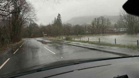 The A593 road near Rothay Bridge. There is an area of green grass to the right which is covered in dark-coloured flood water. The skies are grey and gloomy and it is raining.
