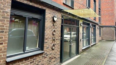 A photo of the exterior of the building which is made from bricks and has black-framed doors and windows