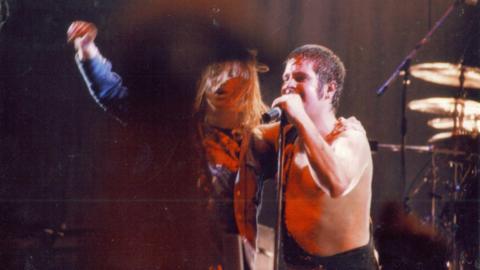 Des Rumney with long blonde hair, has one arm up in the air, and the other hugging Ozzy Osbourne on stage. Ozzy is shirtless as he is singing into a microphone. 