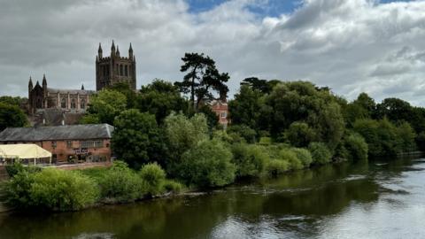 Hereford Cathedral is pictured beside a large river with trees and a smaller red brick building hiding the base of the structure. 