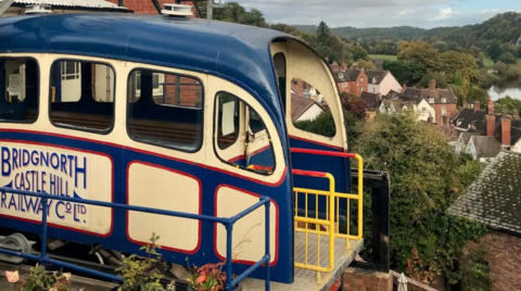 A blue and cream painted cliff railway carriage at the top of a hill with houses and trees below. It is painted with the words Bridgnorth Castle Hill Railway Co Ltd