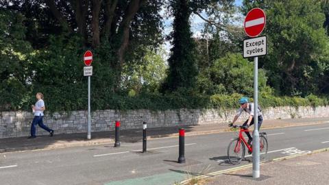 Dryden Road in Exeter with no entry signs in red and white and bollards across the road. A woman is walking on the pavement and a man is cycling on a red bicycle.