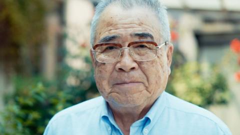 Hiroshima survivor Kenji Nakahara looking into the camera stood outside wearing a blue shirt and glasses with bushes behind him
