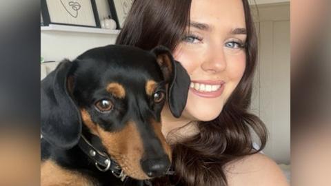 Lucy, who has long brown hair, smiles with Mabel in a selfie. Mabel is a small dachshund with a black coat and tan markings on her face.