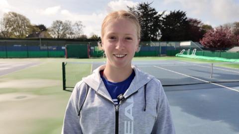 Daniella Britton wearing a grey and black hoodie and a blue t-shirt. She is stood on a blue and green tennis court and is staring at the camera smiling.