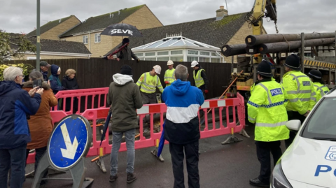 People, including residents and police officers, stand watching workmen preparing to install telecommunications poles.