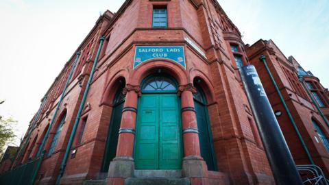 The green door at the entrance to the red brick Salford Lads Club, with two red columns either side. 