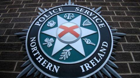 Police Service of Northern Ireland green and black crest against brown brick wall 