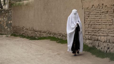 A woman wearing a white burka - a full veil - walking along a road in Afghanistan 