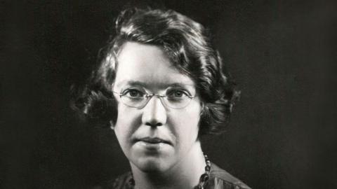 This is a black and white picture. Miss Haining is wearing glasses and has short dark hair. She is wearing a necklace.