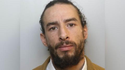 A custody picture of Cody. He has long dark hair pulled back from his face, dark eyebrows and a dark beard. He wears a white collared shirt and what appears to be a tan-coloured jacket. 