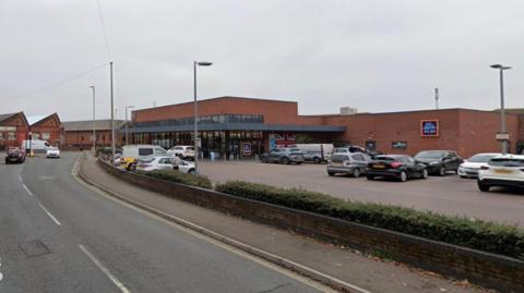 Google Maps street view of an Aldi store on Clifton Road in Gloucester. It is a red brick building with glass panels at the entrance. There is a car park to the side of the store and there are many cars parked up.