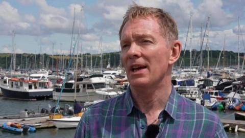 Tourism Minister Sir Chris Bryant wears a green and purple tartan style shirt while stood in a marina in Mylor, Cornwall. There are dozens of boats and yachts behind him.